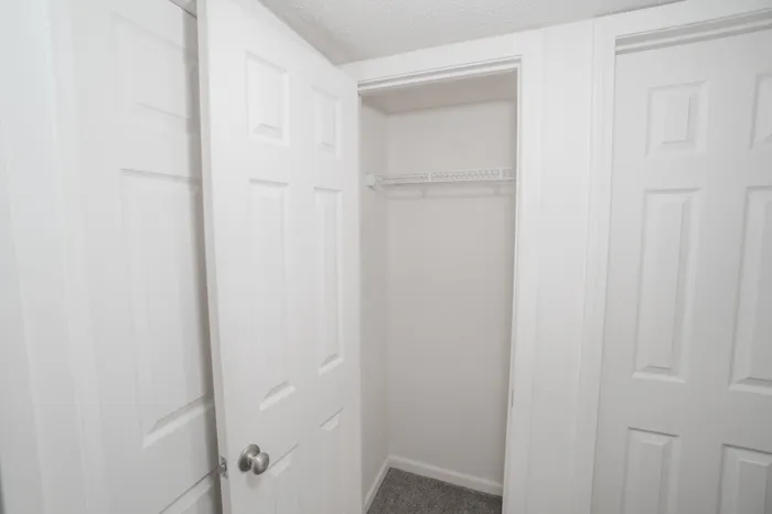 Hallway storage closet at Rosewood Village Apartments featuring dual paneled doors, built in wire shelving, neutral white finishes, and convenient extra storage space.