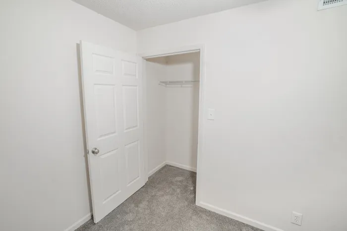 Bedroom closet at Rosewood Village Apartments featuring built in wire shelving, soft gray carpeting, neutral white finishes, and convenient storage space with paneled door access.