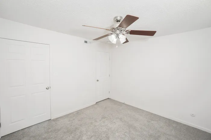Bright bedroom interior at Rosewood Village Apartments with plush gray carpeting, ceiling fan with lighting, neutral paint tones, and spacious layout with closet and hallway access.