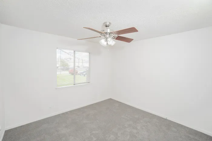 Cozy bedroom at Rosewood Village Apartments featuring soft carpeting, ceiling fan lighting, large window with blinds for natural light, and clean neutral finishes throughout.