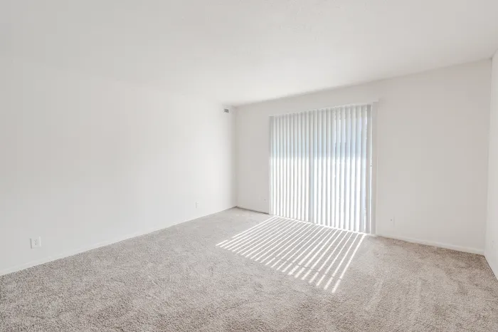 Large, open living room with neutral tones and vertical blinds leading to a private patio or balcony.