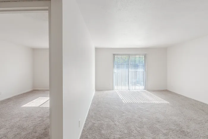 View from the bedroom into a spacious carpeted living room at Rosewood Village Apartments in Springfield, MO, with natural light streaming through vertical blinds.