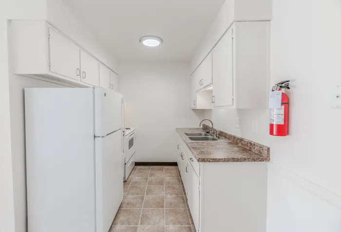 Galley-style kitchen with white appliances, tile flooring, and ample upper and lower cabinet space at Rosewood Village Apartments.