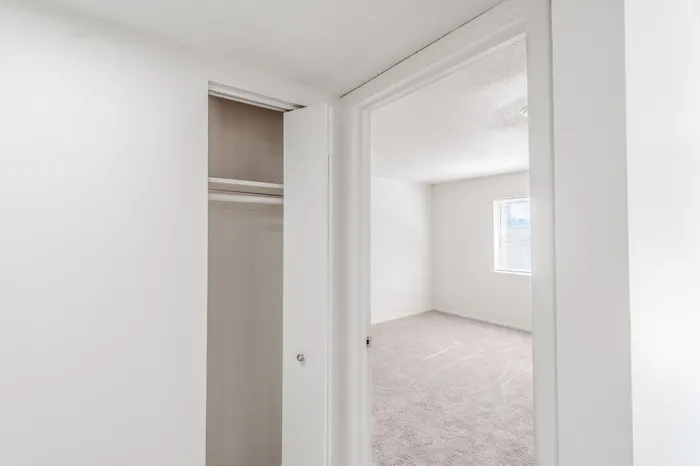 Hallway closet and view into the bedroom featuring a window with blinds and carpeted floors.