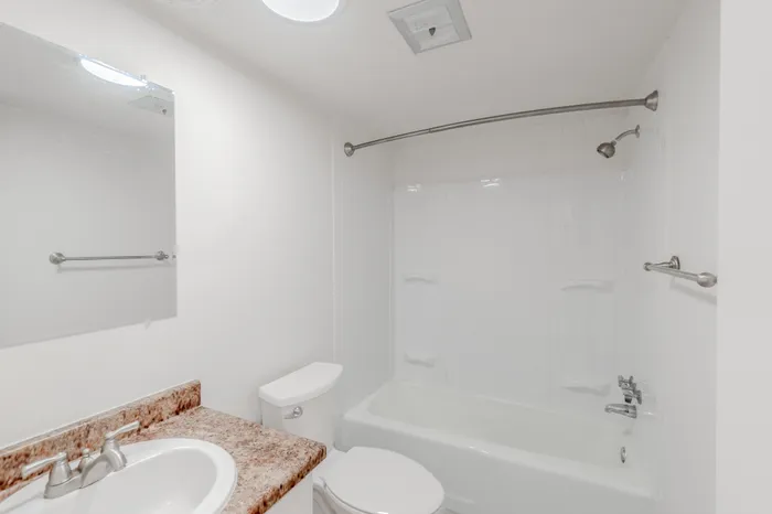 Clean, white bathroom with a full-size tub and shower, large mirror, and marble-style counter.