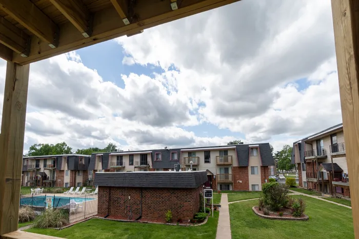 Charming balcony view overlooking a serene apartment complex with lush greenery, inviting walkway, and sparkling pool under a vibrant sky.