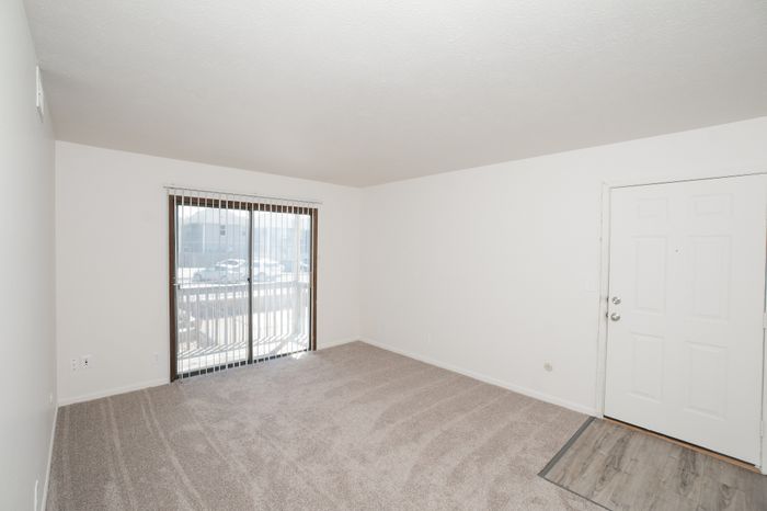 Spacious living room at El Chaparral Apartments with carpet, entry door, sliding glass door, and neutral finishes.