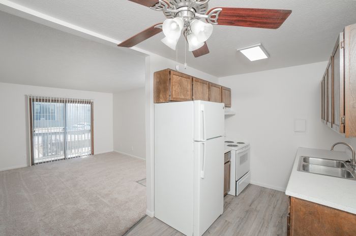 Classic kitchen and living area at El Chaparral Apartments with wood cabinets, white appliances, ceiling fan, carpet, and sliding glass door.