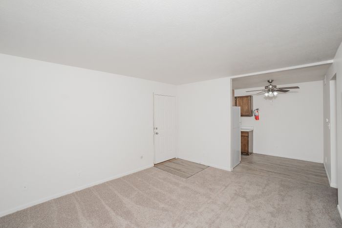 Open living room at El Chaparral Apartments with carpet, white walls, entry door, and view into the classic kitchen.