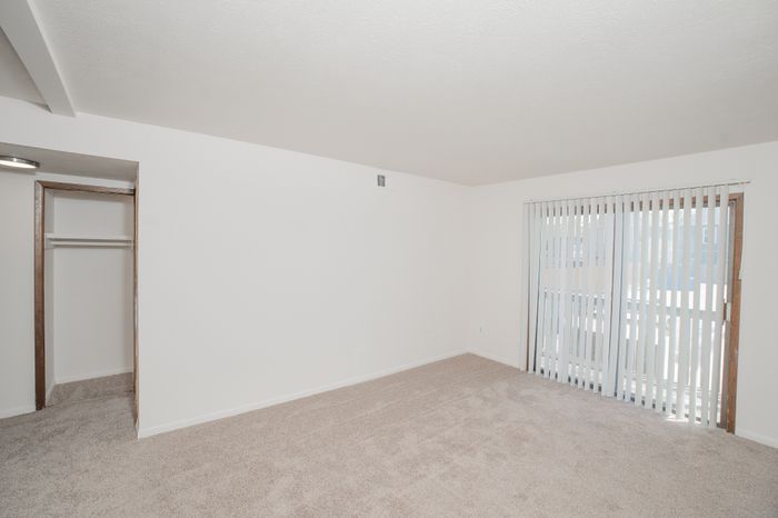 Carpeted living room at El Chaparral Apartments with sliding glass door, vertical blinds, closet, and white walls.