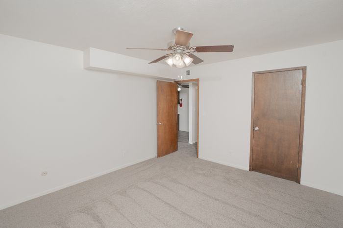 Carpeted bedroom at El Chaparral Apartments with ceiling fan, white walls, closet access, and neutral finishes.