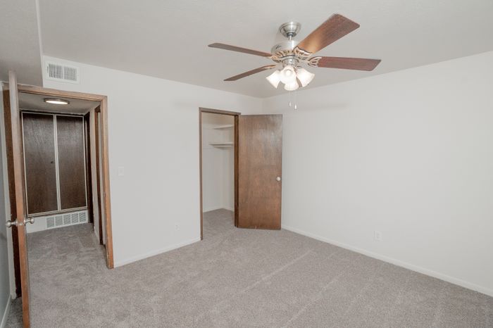 Bedroom at El Chaparral Apartments with carpet, ceiling fan, window with blinds, and white walls.
