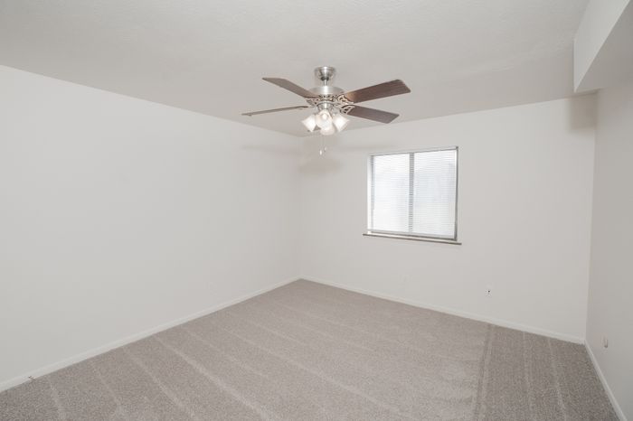 Spacious carpeted bedroom at El Chaparral Apartments with ceiling fan, window with blinds, and white walls.