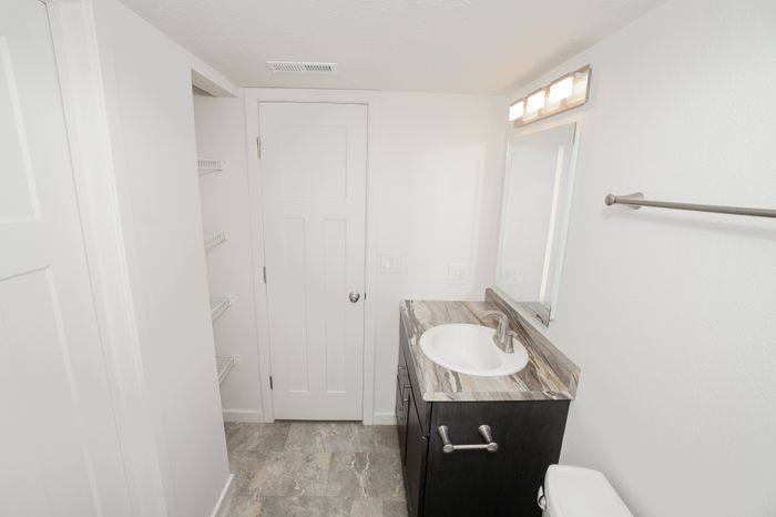 Renovated bathroom at El Chaparral Apartments with gray tile style flooring, a dark vanity, marble look countertop, white sink, mirror, and linen shelving.