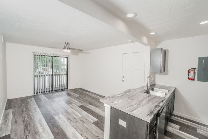 Open living and kitchen area at El Chaparral Apartments with gray wood style flooring, ceiling fan, sliding glass door, and updated kitchen finishes.