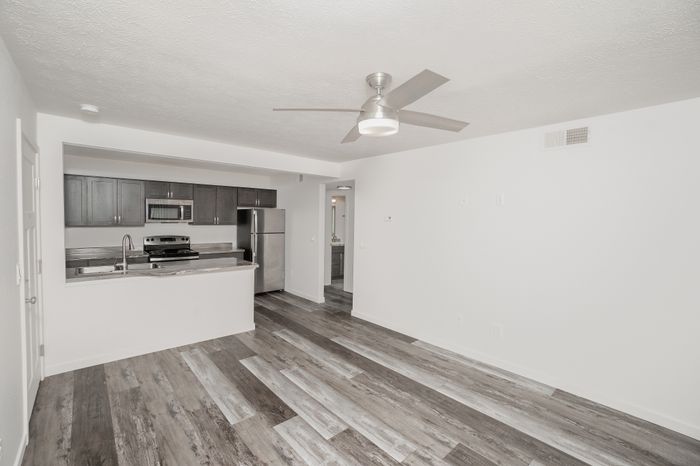 Spacious renovated living room at El Chaparral Apartments with gray wood style flooring, ceiling fan, open kitchen, and bright white walls.