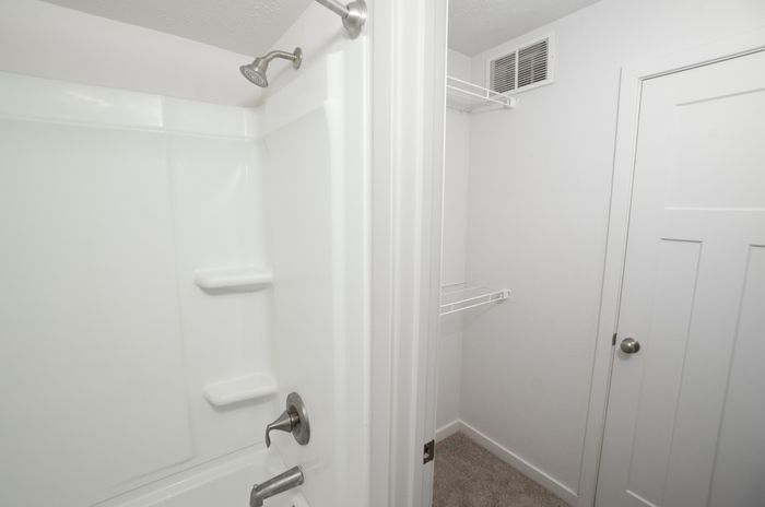 Bathroom and closet area at El Chaparral Apartments with a white tub and shower, wire shelving, carpet, and white paneled door.