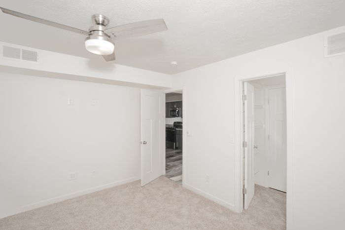 Carpeted bedroom at El Chaparral Apartments with white walls, ceiling fan, closet access, and a view into the updated kitchen.