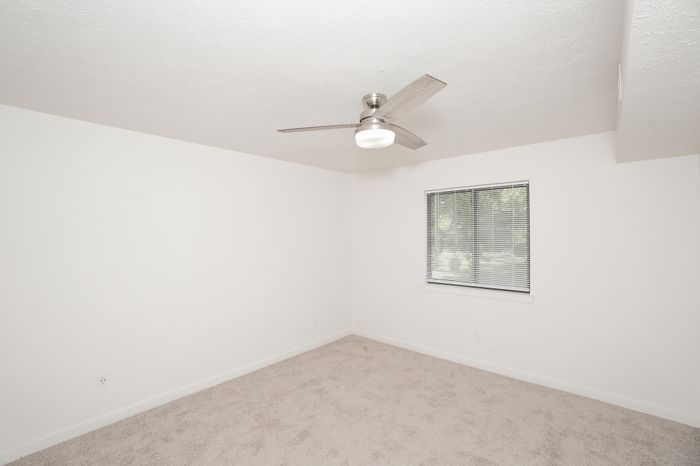 Spacious bedroom at El Chaparral Apartments with soft carpet, white walls, ceiling fan, and window with blinds.