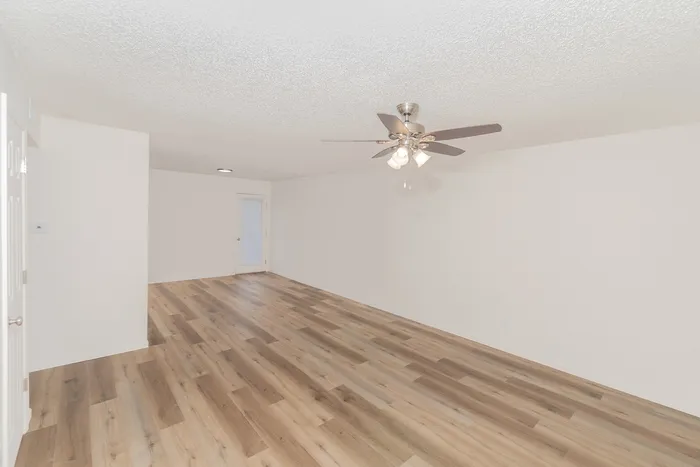 Bright and open one bedroom apartment living room at Quail Ridge East Apartments featuring wood style flooring, white walls, ceiling fan with light fixture, and private patio door with blinds.