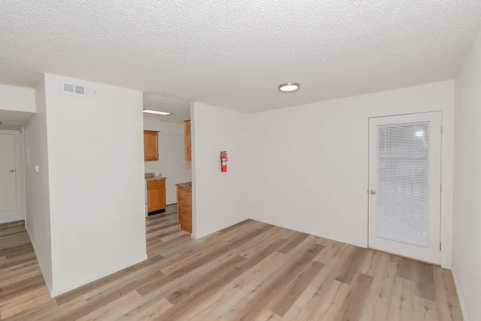 Open dining area and entry to kitchen in a one bedroom apartment at Quail Ridge East Apartments featuring updated flooring, white walls, natural light, and patio access door with blinds.
