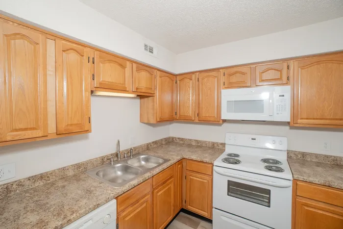 Compact apartment kitchen at Quail Ridge East Apartments with oak cabinetry, laminate countertops, white appliances, double sink, microwave, and wood style flooring.