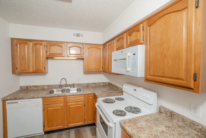 Updated kitchen interior at Quail Ridge East Apartments featuring oak cabinets, white electric stove and microwave, laminate countertops, dishwasher, and bright overhead lighting.