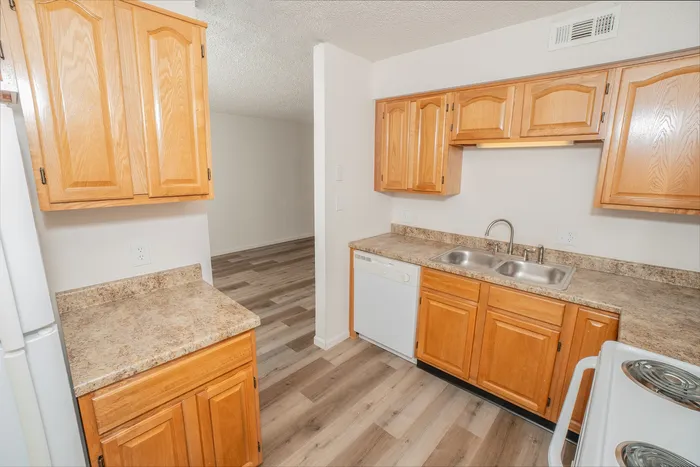 Bright apartment kitchen at Quail Ridge East Apartments with oak cabinets, laminate countertops, double sink, dishwasher, and wood style flooring leading into the living area.