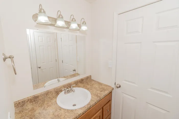 Updated bathroom vanity at Quail Ridge East Apartments featuring laminate countertops, wood cabinetry, brushed nickel fixtures, large mirror, and modern four light vanity fixture.