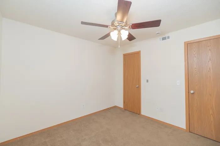 Bright and inviting room with warm carpet flooring, dual wooden doors, and a modern ceiling fan.