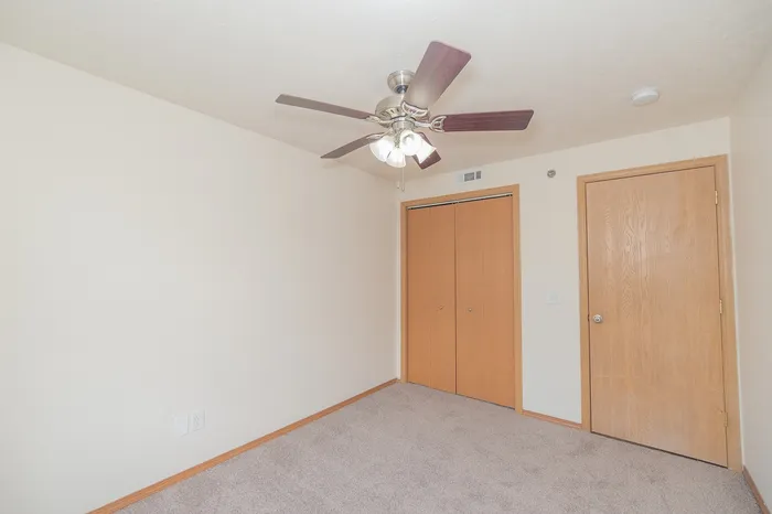 Bright, minimalistic bedroom featuring soft carpet flooring, a sleek ceiling fan, and ample closet space.