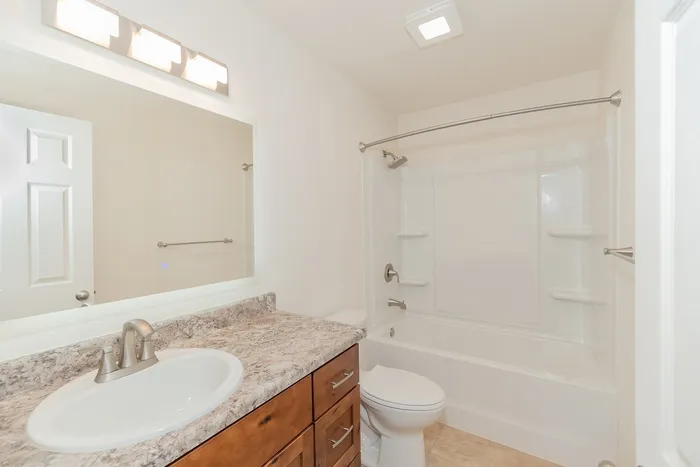 Renovated bathroom at Chesterfield Village with a wood vanity, stone look countertop, large mirror, and white tub and shower.