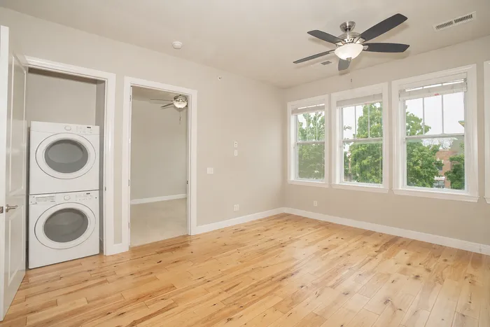 Bright and inviting room with hardwood floors, a ceiling fan, large windows offering lush green views, and an in-unit washer and dryer. Perfect for modern living.
