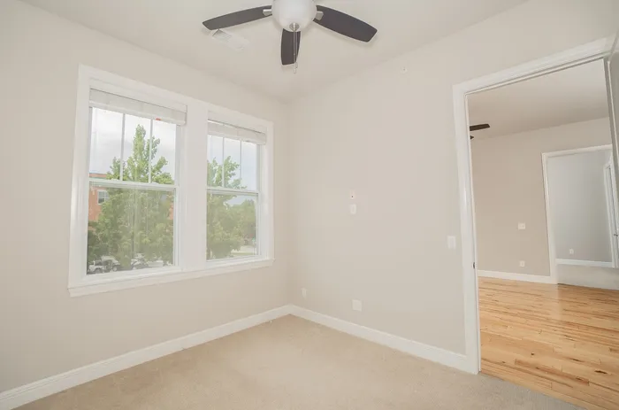 Bright room with large windows showcasing a serene view, featuring modern ceiling fan, plush carpet, and elegant hardwood flooring transition.