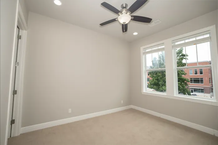 Bright and inviting empty room with modern ceiling fan, large windows providing natural light, and a view of surrounding buildings.