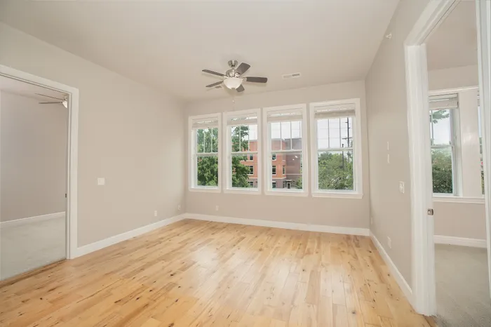 Bright apartment room with elegant wood flooring, large windows offering lush green views, and a sleek ceiling fan.
