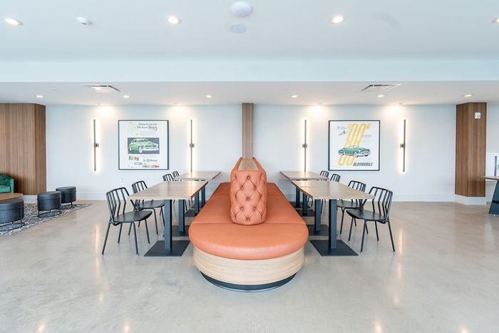 Modern communal area featuring sleek tables, chic black chairs, and a stylish orange cushioned bench against a backdrop of vintage car posters.