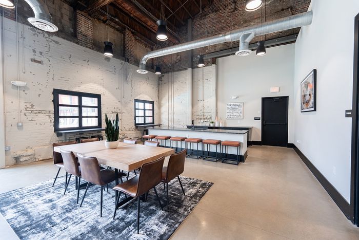 Modern loft-style dining and kitchen area with exposed brick, high ceilings, sleek table, and leather chairs.
