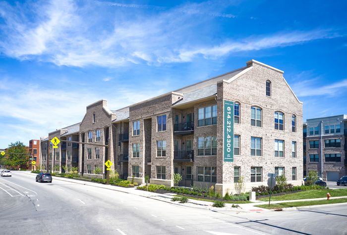 Elegant brick apartment building with spacious balconies, surrounded by lush landscaping and clear blue skies.