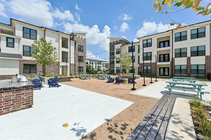 Apartment courtyard with gravel on the ground, a firepit with four blue lawn chairs, and light blue picnic benches