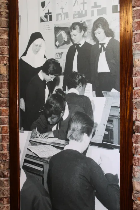 Black and white historical photograph of students diligently working on art projects under the guidance of a nun and teachers in a classroom setting.