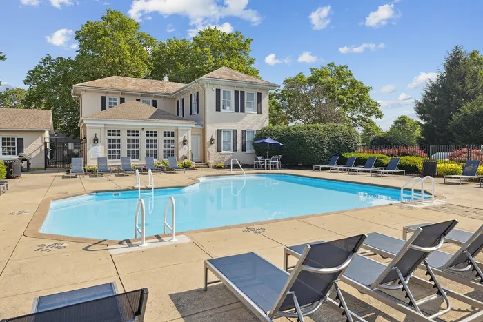 Inviting swimming pool area with elegant loungers and a charming house surrounded by lush greenery under a clear blue sky.