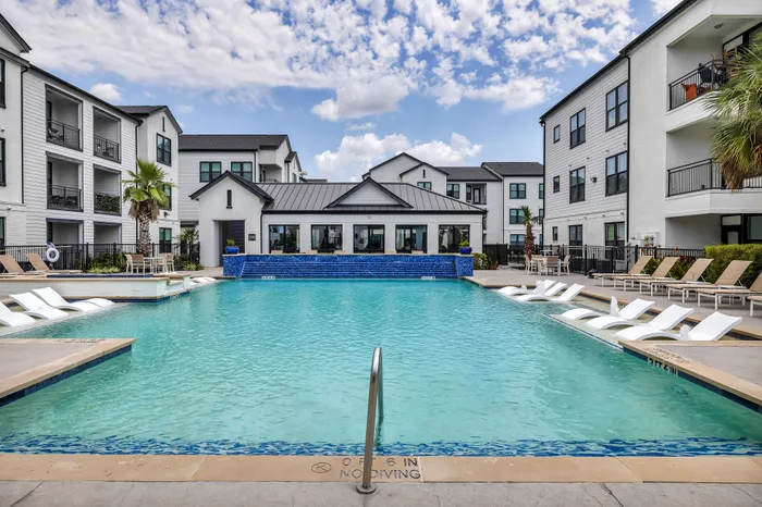 Luxurious apartment complex featuring a stunning outdoor pool area with sleek lounge chairs, vibrant blue accents, and modern architecture against a backdrop of clear skies.