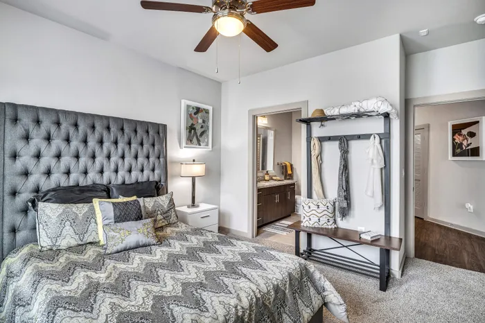 Elegant bedroom with a plush gray headboard, stylish bedding, and a ceiling fan, leading to a modern en-suite bathroom.