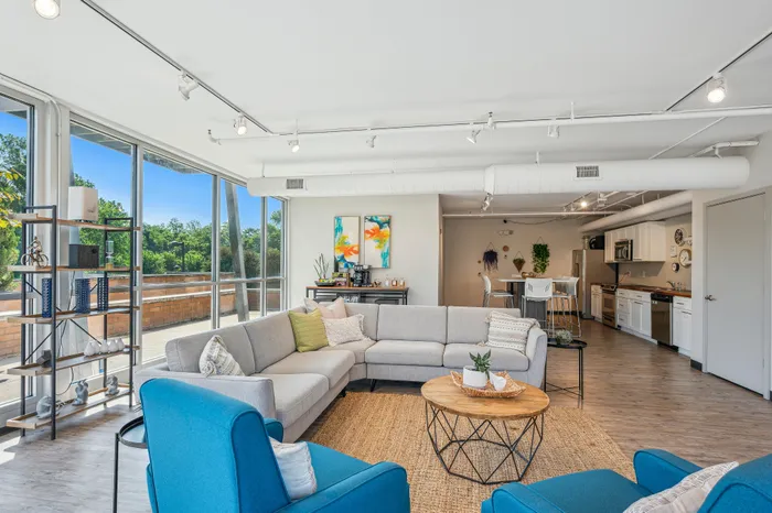 Bright and modern living space with panoramic windows, featuring a cozy L-shaped sofa, blue armchairs, and a chic open kitchen perfect for entertaining.