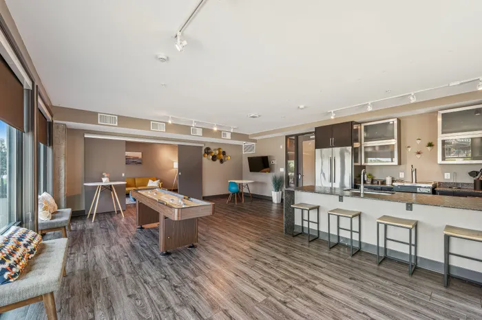 Spacious and modern entertainment room featuring sleek hardwood floors, a stylish kitchenette with bar seating, and a cozy lounge area with ample sunlight.