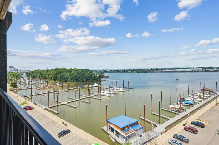 Scenic waterfront view from a balcony overlooking a marina with boats and lush trees under a bright blue sky.