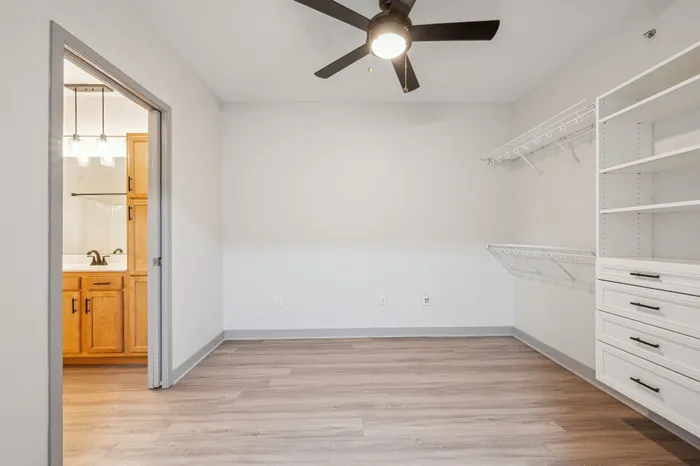 Bright and modern room featuring sleek hardwood floors, a ceiling fan, and spacious built-in shelving, with a view into a stylish en-suite bathroom.