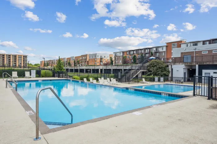 Modern apartment complex featuring a pristine outdoor pool with lounge chairs, surrounded by lush greenery and stylish multi-story buildings under a clear blue sky.