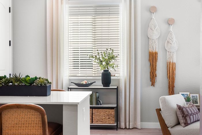 Modern living space with elegant macrame wall hangings, natural light filtering through blinds, and a stylish seating area accented by a vase with greenery.